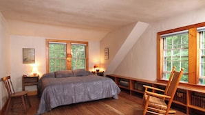 Een slaapkamer met een houten vloer, een bed met een grijs bloemenbeddengoed, twee nachtkastjes met lampen, een schommelstoel en een raam met uitzicht op groen.