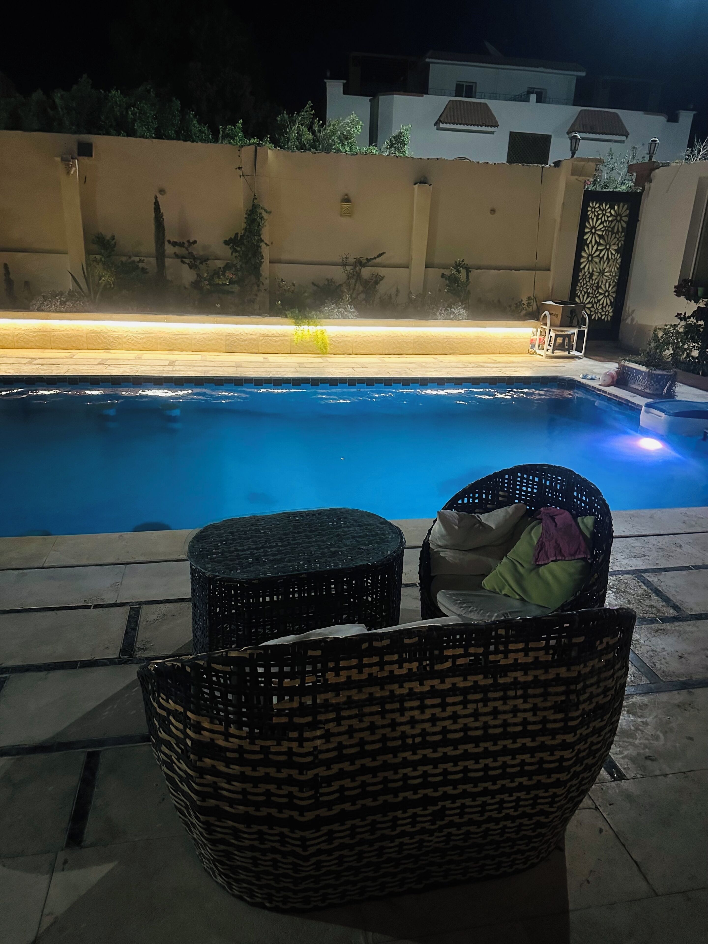 A swimming pool with two wicker chairs and a small table, surrounded by a low wall and a potted plant.