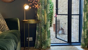 Een kamer met een groene bank, een klein rond tafeltje met een lamp, een spiegel en een glazen schuifdeur naar een buitenruimte met een bakstenen muur en begroeiing.
