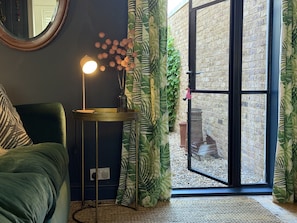 A room with a green sofa, a small round table with a lamp, a mirror, and a sliding glass door leading to an outdoor area with a brick wall and greenery.