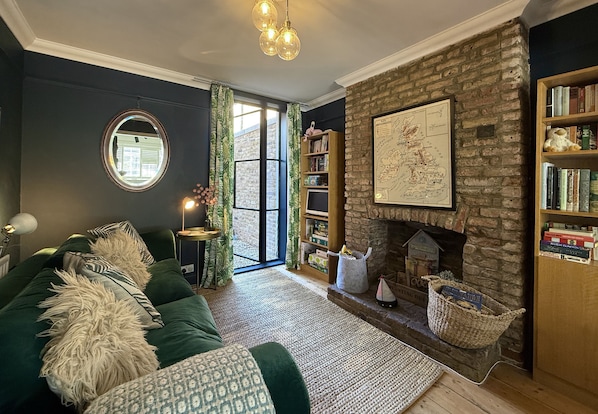 A cozy living room with a green sofa, a fireplace, a bookshelf, and a large window with curtains.