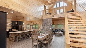 A spacious wooden cabin interior with a central dining area, kitchen, and a loft bedroom with a wooden staircase. - Stargazer Chalet 5 Bedroom Cabin by RedAwning (Sevierville)