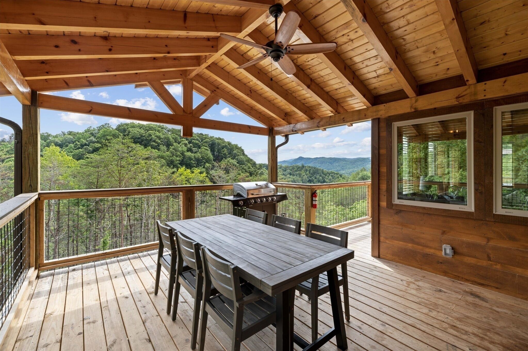 Une terrasse en bois avec une table et des chaises, un ventilateur de plafond et une vue sur une chaîne de montagnes boisée.