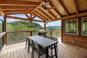 Eine Holzdielen-Terrasse mit Tisch und Stühlen, einem Deckenventilator und Blick auf einen bewaldeten Gebirgszug.