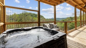 A hot tub area with a wooden deck, a cushioned seating area, and a view of a forested landscape. - Stargazer Chalet 5 Bedroom Cabin by RedAwning (Sevierville)