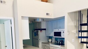 A compact kitchen with white cabinets, a microwave, and a refrigerator. There is a small island with stools and a door leading to another room.