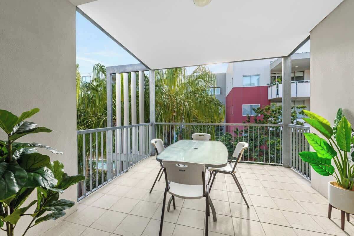 A balcony with a table and chairs, surrounded by potted plants and a view of buildings and greenery.