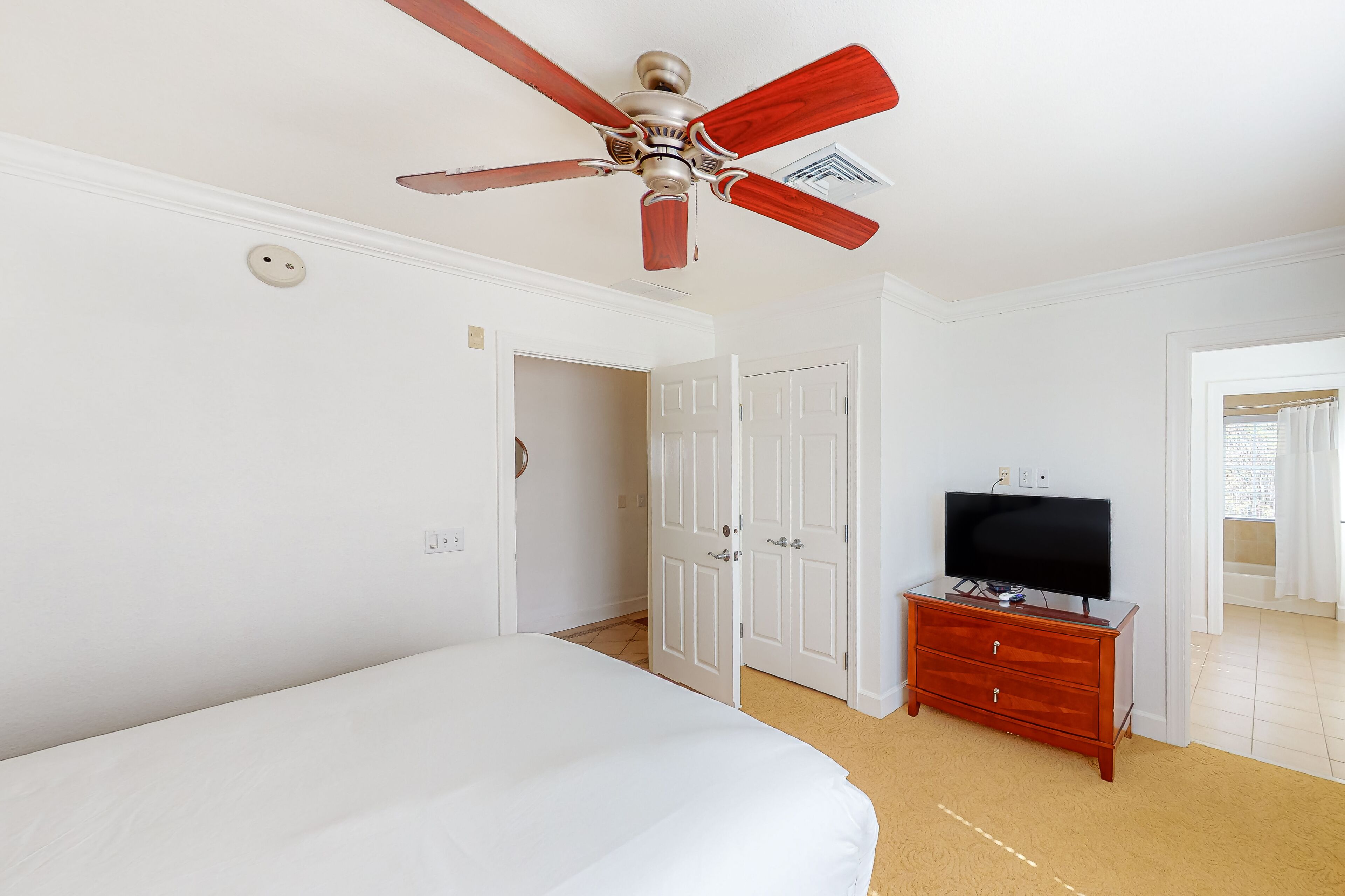 Ein Schlafzimmer mit einem Bett, einer Holzkommode, einem Fernseher, einem Deckenventilator und weißen Türen.