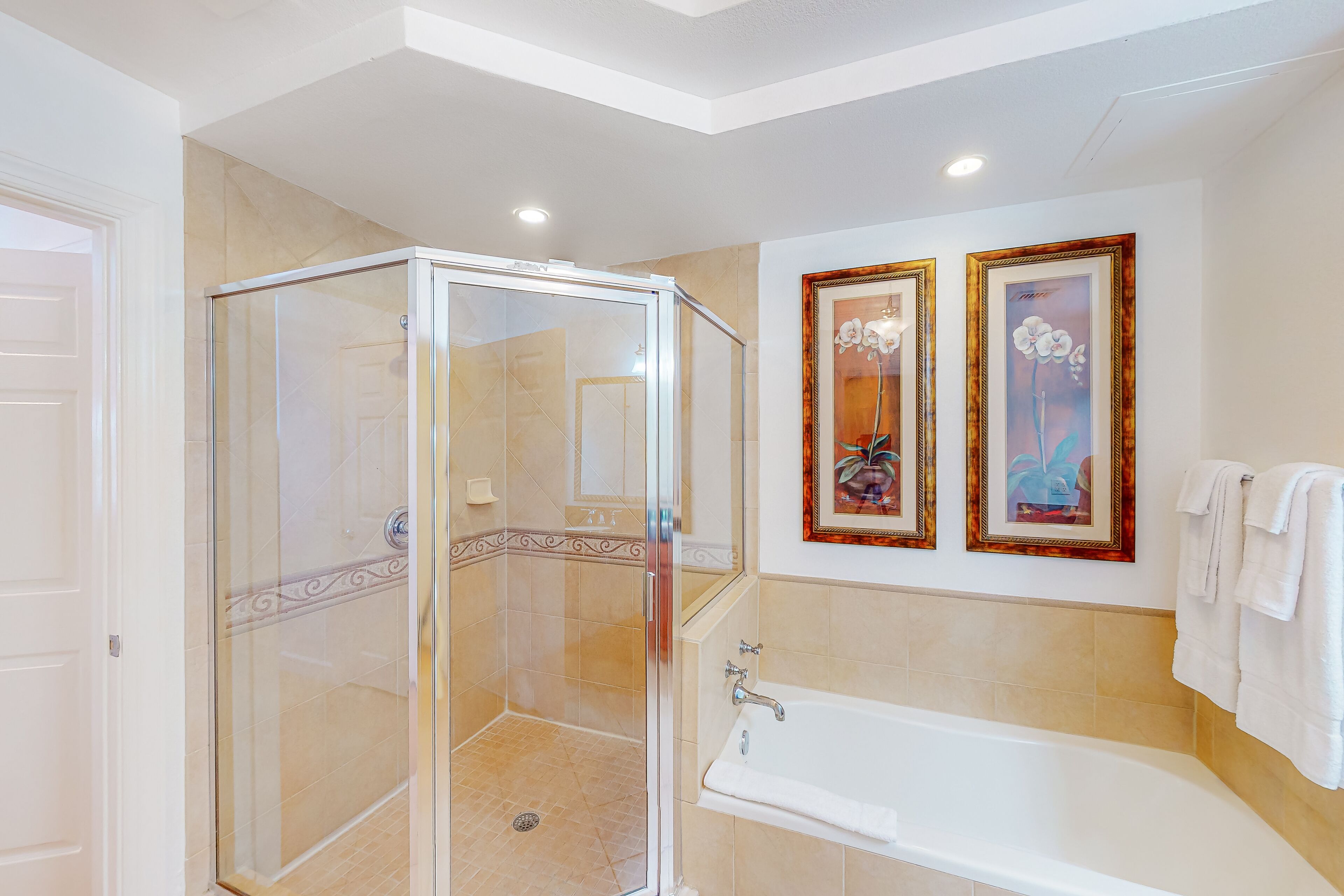 A modern bathroom with a glass-enclosed shower, a white bathtub, and two framed floral paintings on the wall.