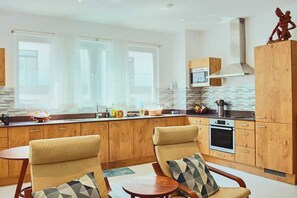 A modern kitchen with wooden cabinets, a built-in oven, and a microwave. There are two chairs with cushions and a small table.