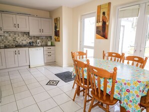 A kitchen with white cabinets, a dishwasher, and a tiled floor. There is a dining table with chairs and a floral tablecloth.