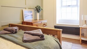 A bedroom with a bed, a wooden dresser, a radiator, and a window with blinds. - Gwel Y Môr (Beaumaris)