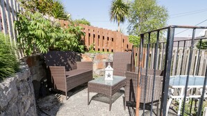 An outdoor patio with wicker furniture and a glass table, surrounded by a wooden fence and greenery. - Gwel Y Môr (Beaumaris)