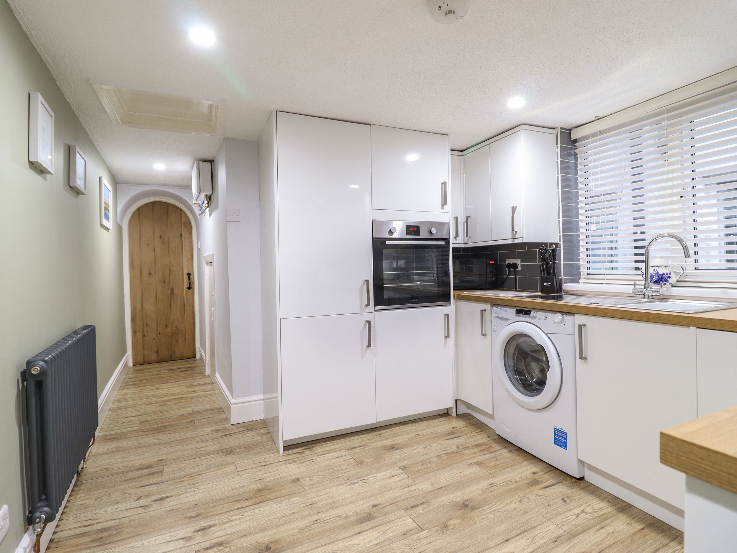 A modern kitchen with white cabinets, a built-in oven, a washing machine, and a window with blinds.