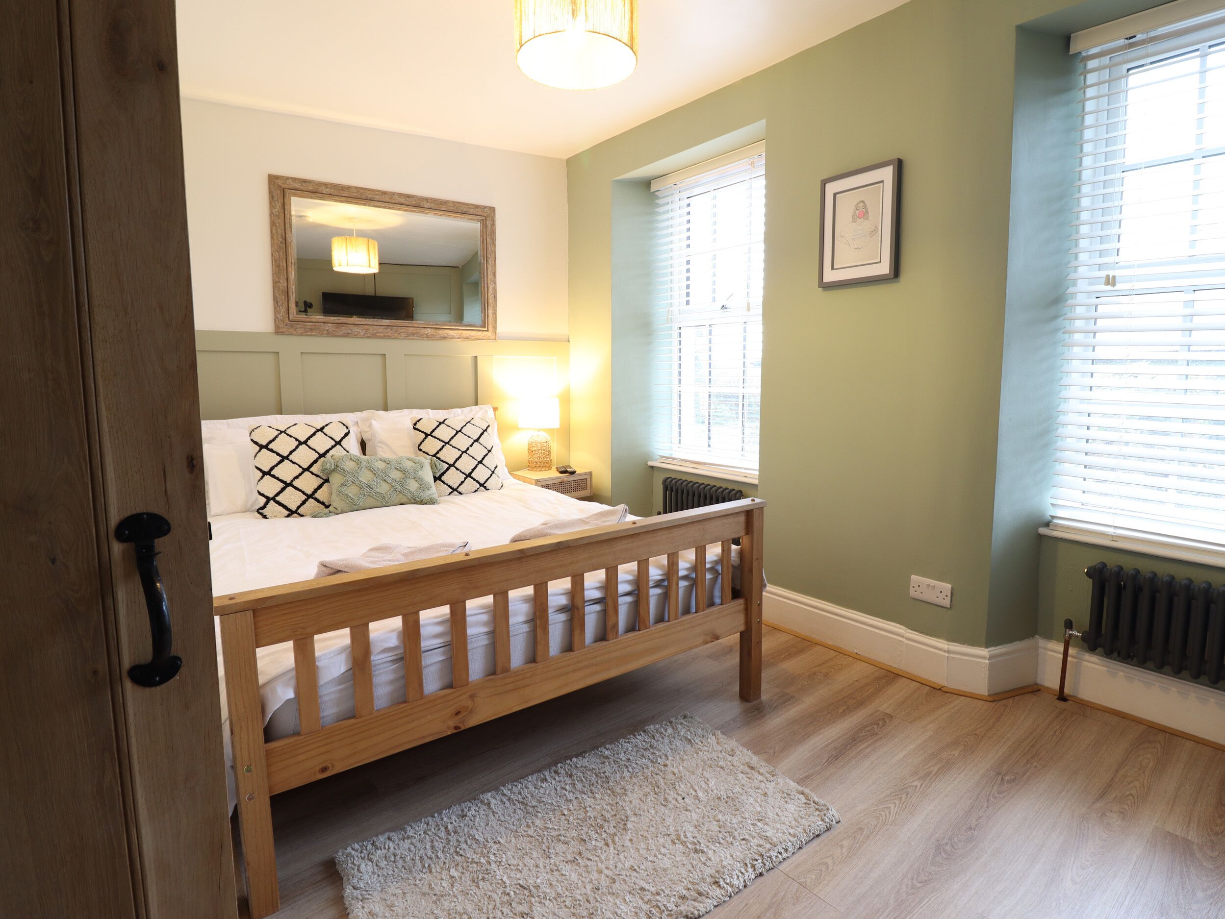 A bedroom with a wooden bed, a bedside table with a lamp, a radiator, and a window with blinds.