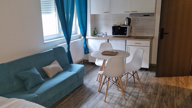 Ein modernes Schlafzimmer mit Bett, blauem Sofa, Esstisch mit Stühlen, Küchenzeile inklusive Mikrowelle und Holztür.