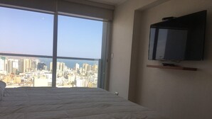 A bedroom with a large bed, a city view through a window, and a mounted television on the wall. - Miraflores Upper Pardo Apartment 3bedroom/4baths (Miraflores)