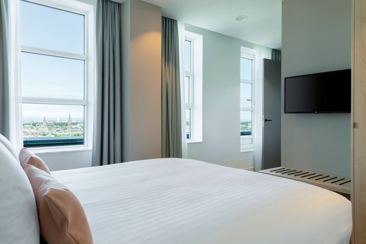 A hotel room with a large bed, a television mounted on the wall, and a window offering a city view.
