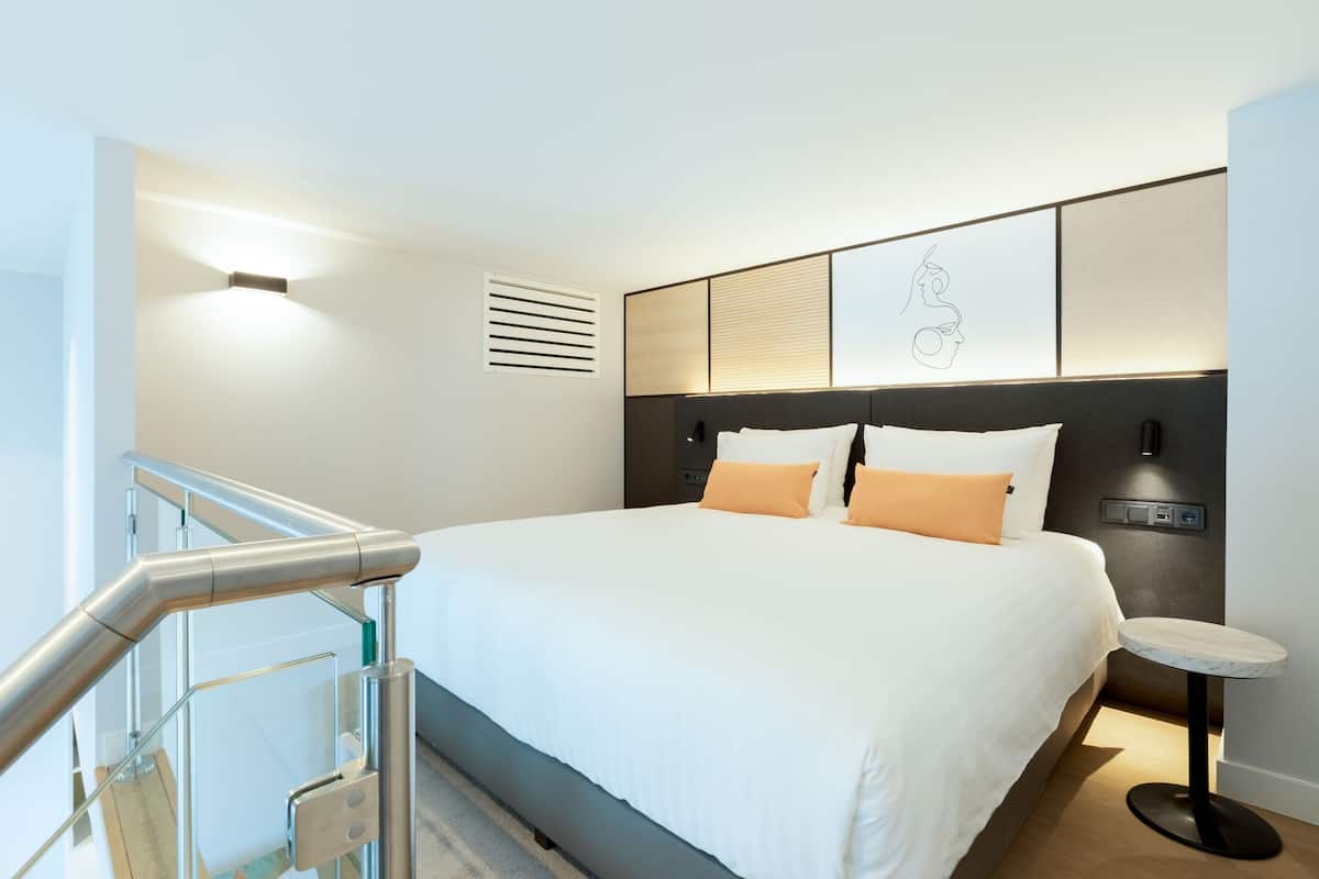 A modern hotel room with a large bed, a small round table, and a staircase with glass railings.