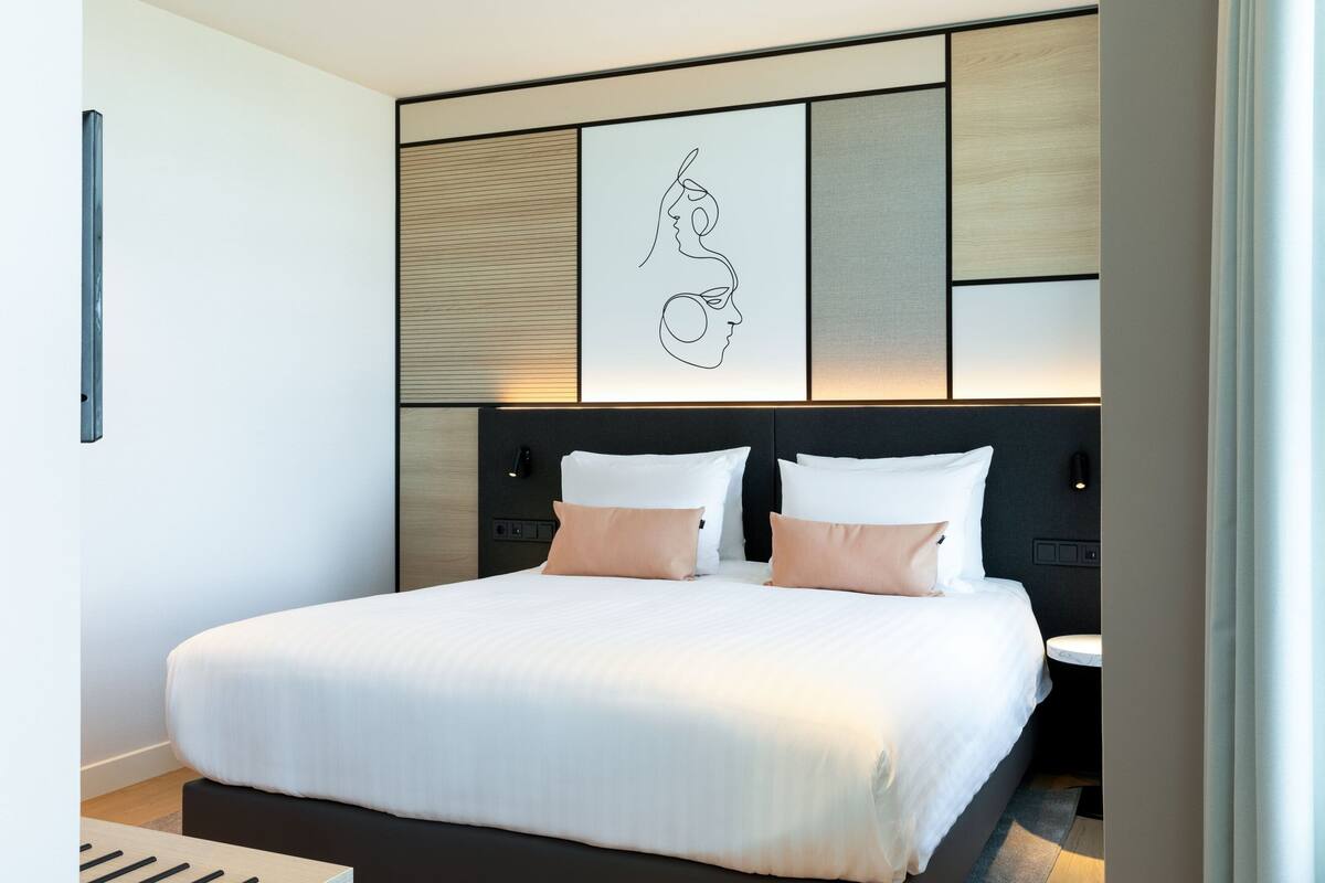 A modern hotel room with a large bed, a headboard with a geometric design, and a wall-mounted artwork.