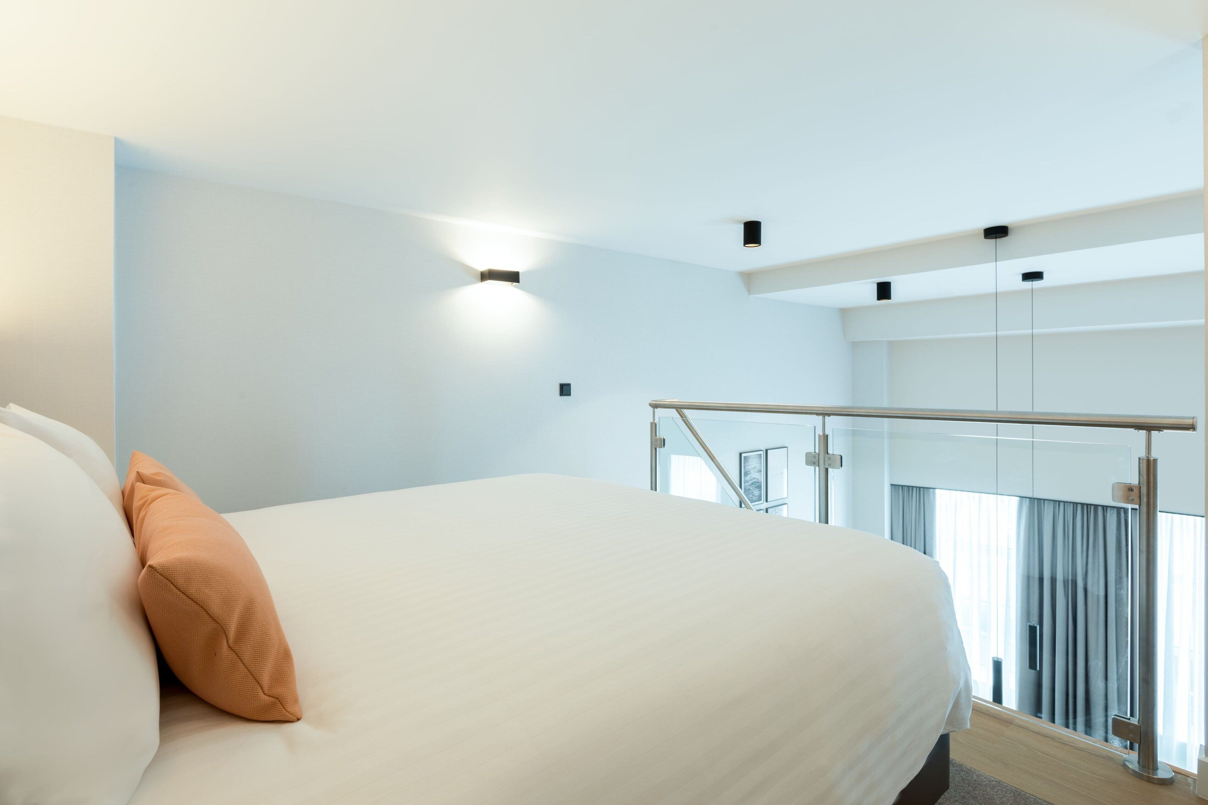 Una habitación de hotel moderna con una cama grande, un balcón con barandilla de vidrio y vista a la ciudad.