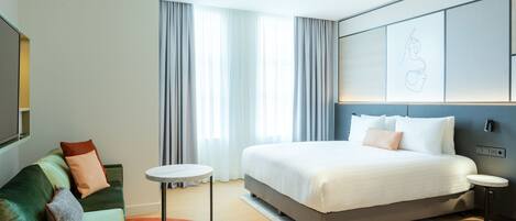 A modern hotel room with a large bed, a green sofa, a small round table, and a wall-mounted artwork.
