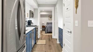 Um corredor estreito com uma cozinha de um lado, uma cama com colcha cinza ao centro e uma porta com um detalhe de folha na parede.