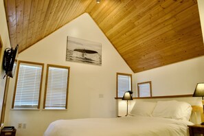 A bedroom with a sloped wooden ceiling, two windows with blinds, a bed with white linens, a lamp on the nightstand, and a wall-mounted artwork of a whale. - Bright & Beautifully Refreshed Cottage - 2 min from Ocean! (Ucluelet)