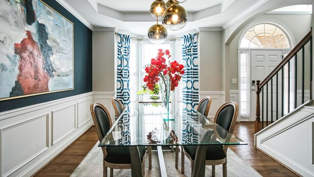 Un comedor con mesa de vidrio, sillas, candelabro, un cuadro grande y una escalera.