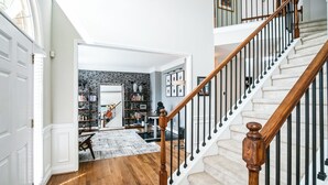 Un pasillo amplio con suelo de madera, una escalera con barandilla de madera y una puerta blanca a la izquierda.