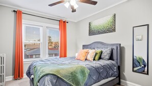 Una camera da letto con un letto, una ventola a soffitto, una finestra con tende e un quadro appeso al muro.