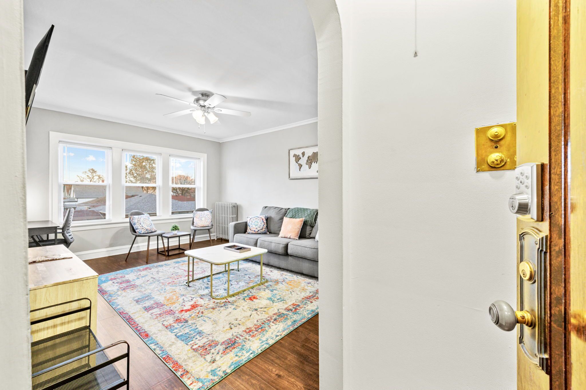 A modern living room with a sofa, coffee table, and chairs. There is a ceiling fan, a window with blinds, and a door with a brass handle.