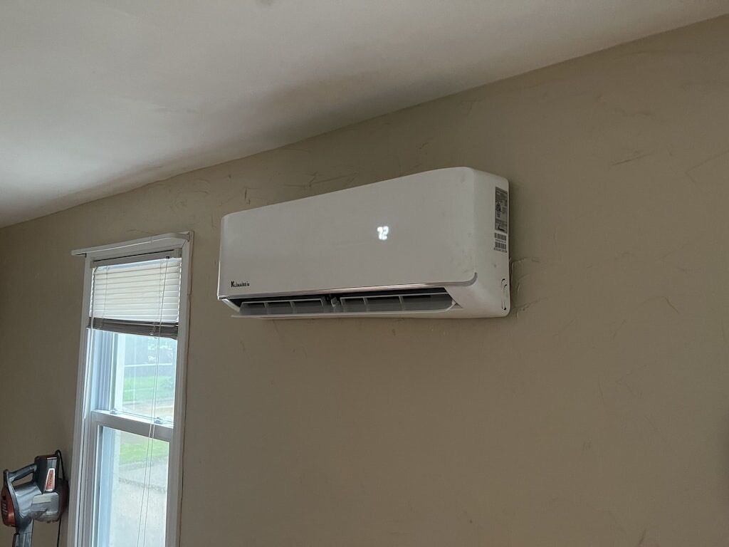 A white air conditioning unit mounted on a beige wall, a window with blinds, and a power tool on the floor.