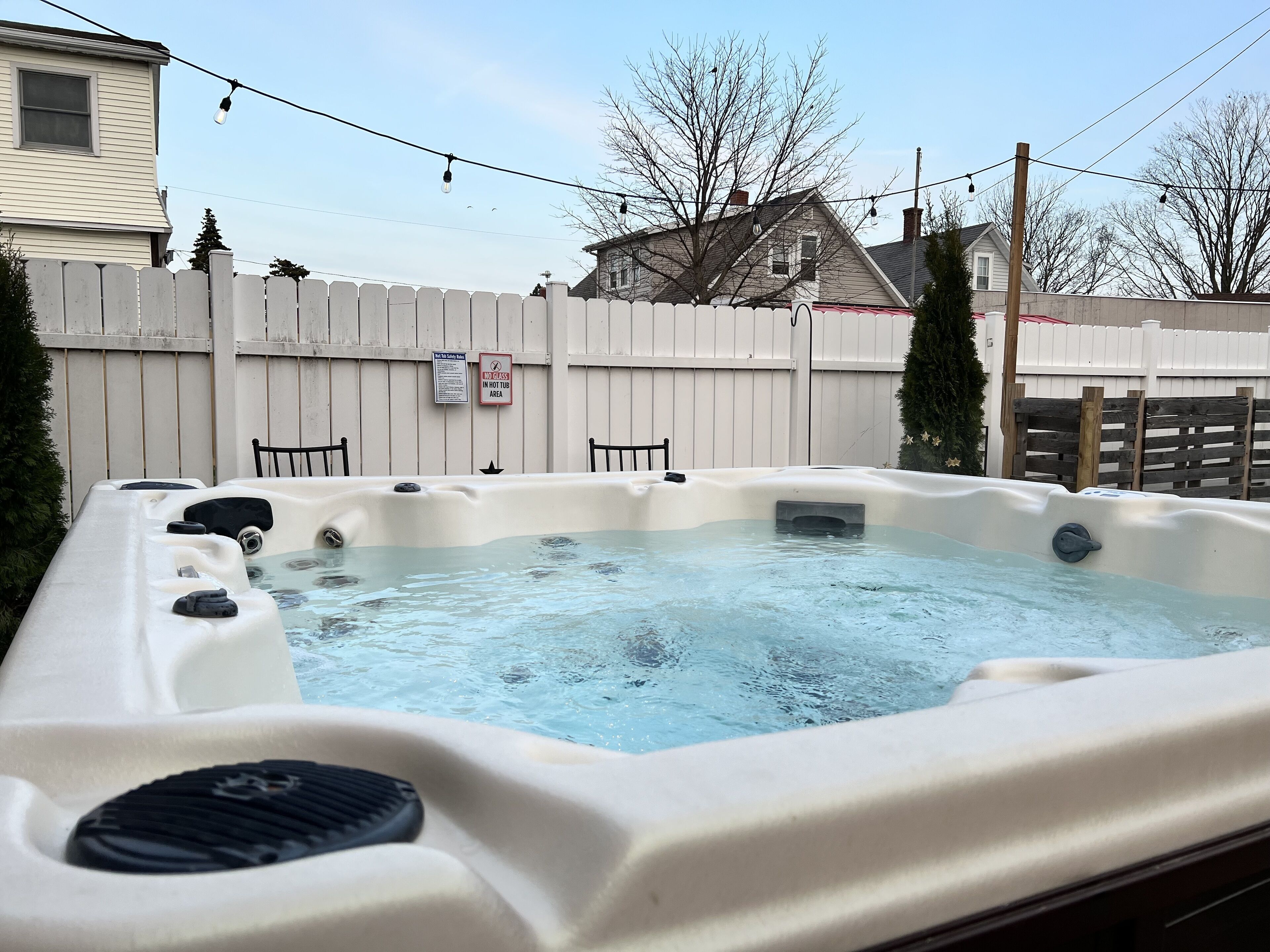 Un jacuzzi en un patio trasero con valla de madera, un letrero que indica la prohibición de usar el jacuzzi en el área del jacuzzi, y una casa al fondo.