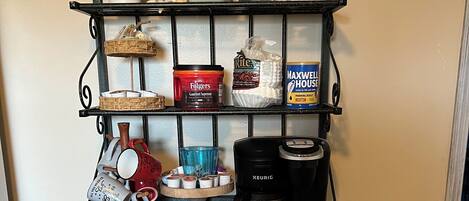 Una encimera de cocina con una cafetera, tazas, un expositor para pasteles y varios utensilios de cocina.