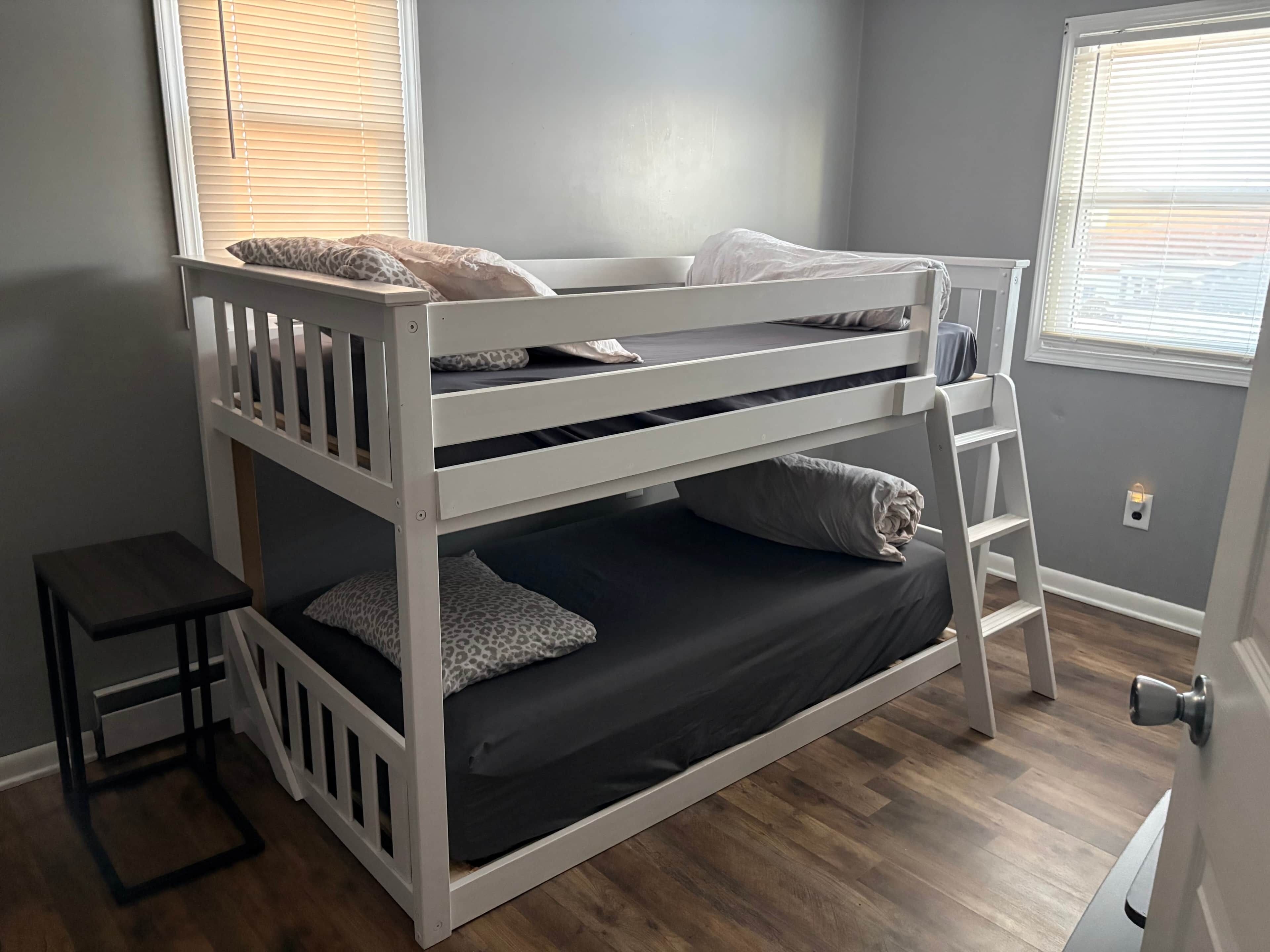 A bunk bed with a ladder, a small table, and a window with blinds.