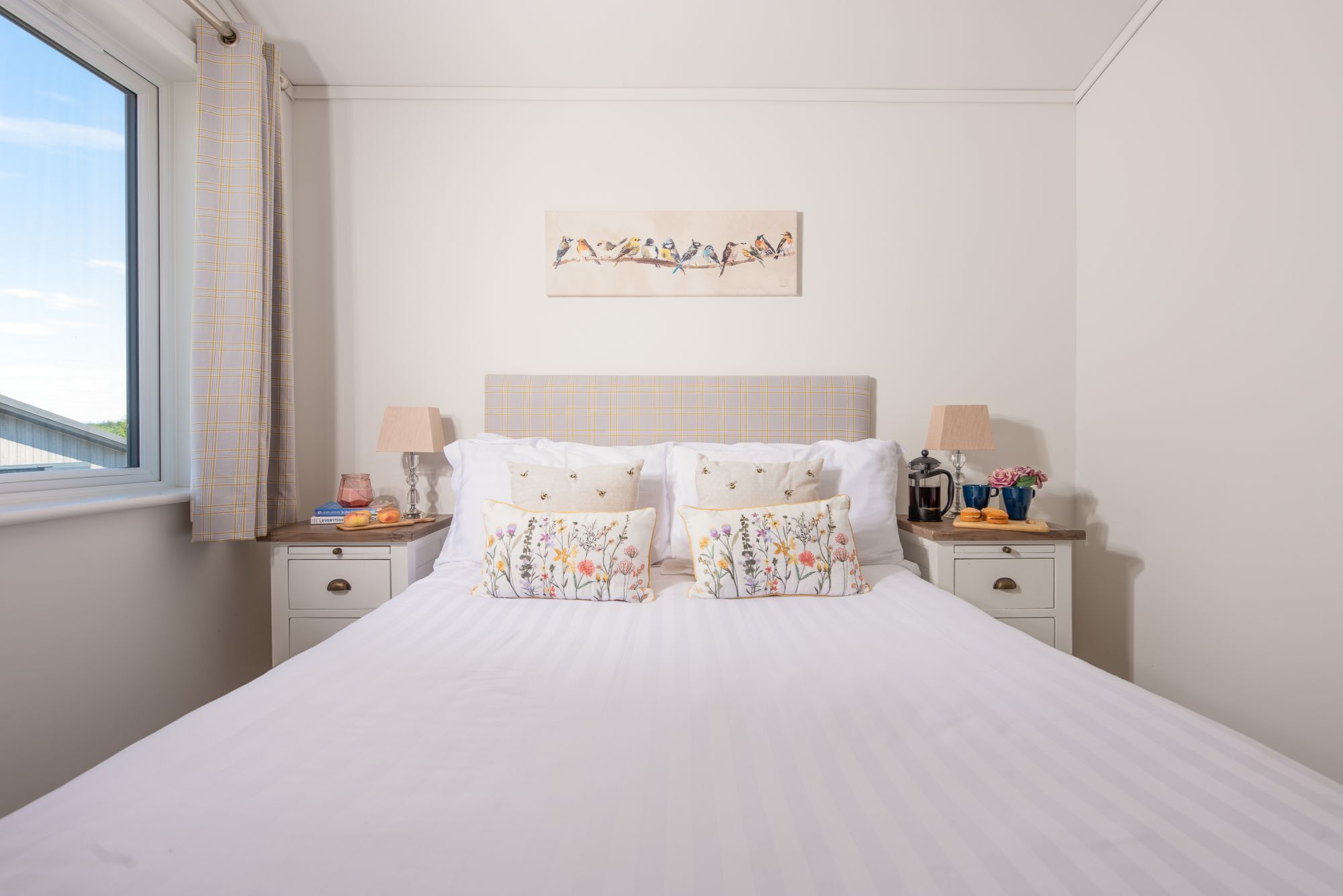 A bedroom with a large bed, two bedside tables, a window with curtains, a headboard, and a wall-mounted painting.