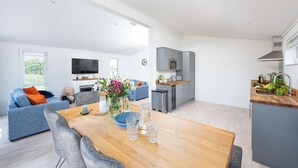 A modern living room with a wooden dining table, grey sofas, and a kitchen area with grey cabinets and a built-in oven.