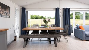 A dining area with a wooden table, chairs, and a bench. There is a view of a field through large windows.