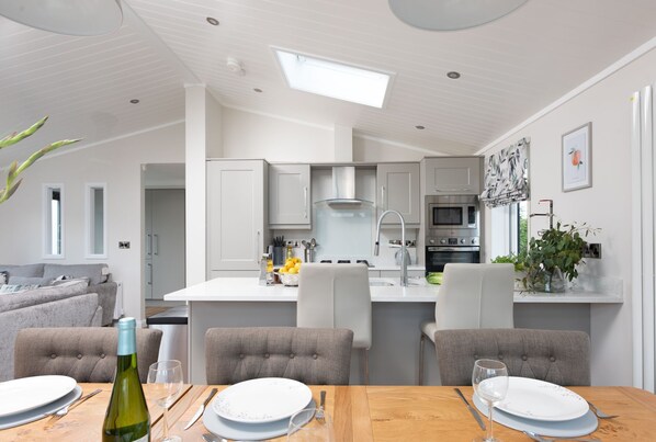 A modern kitchen with a central island, grey upholstered chairs, and a dining table set for two. - Orchid Lodge, 23 Roadford Lake Lodges (Lifton)