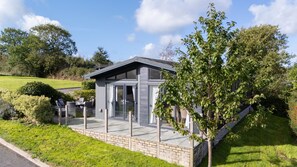 A modern building with a glass door entrance, a patio area with outdoor furniture, and a well-maintained lawn. - Orchid Lodge, 23 Roadford Lake Lodges (Lifton)