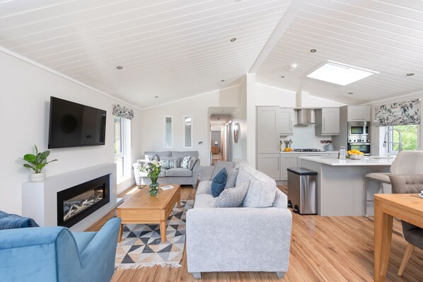 A modern living room with a sofa, coffee table, and TV. A kitchen with a dining area is visible in the background. - Orchid Lodge, 23 Roadford Lake Lodges (Lifton)