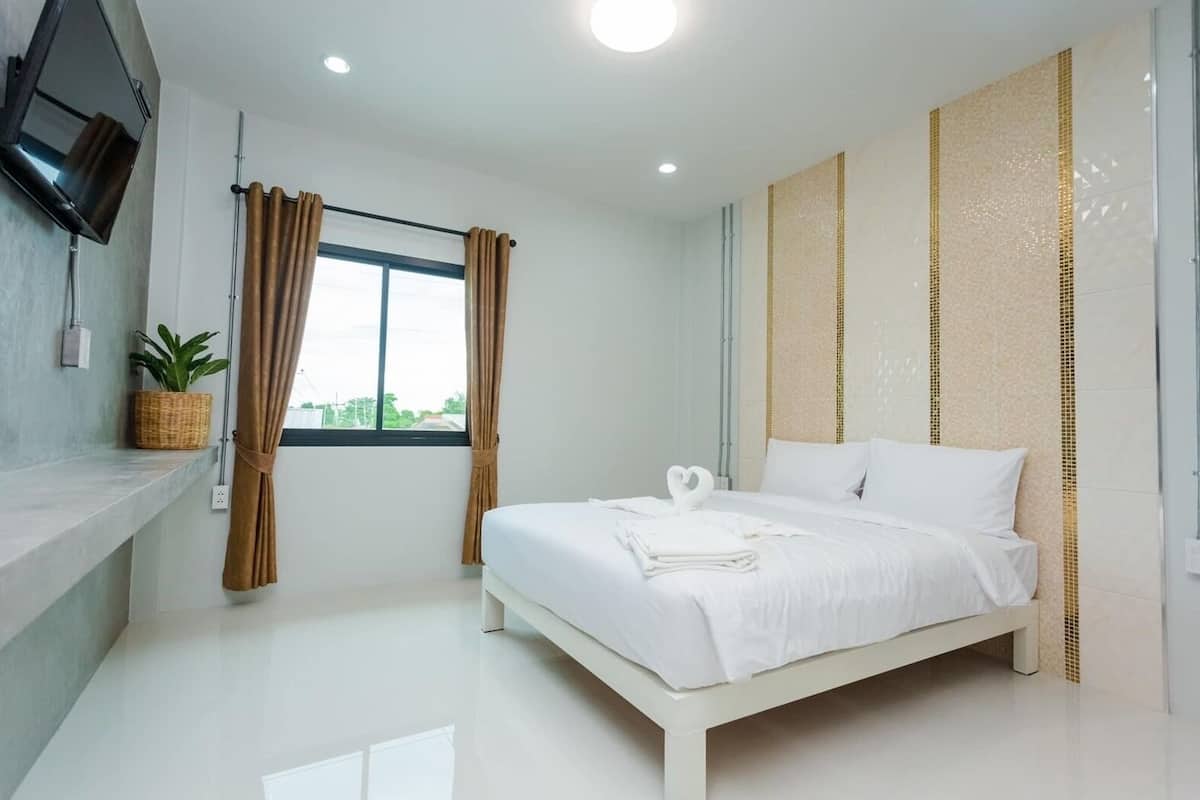 A modern hotel room with a large bed, a flat-screen TV mounted on the wall, a window with curtains, and a potted plant on a shelf.