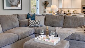 Un salon moderne avec un canapé d’angle gris, un plateau blanc sur lequel se trouvent une bouteille et des verres, et un vase décoratif.