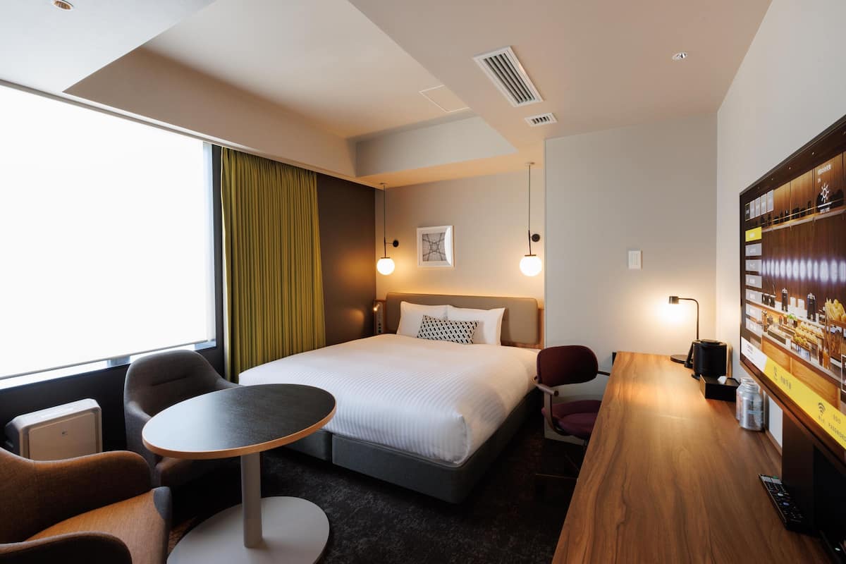 A hotel room with a bed, a desk, a chair, a small table, and a minibar.