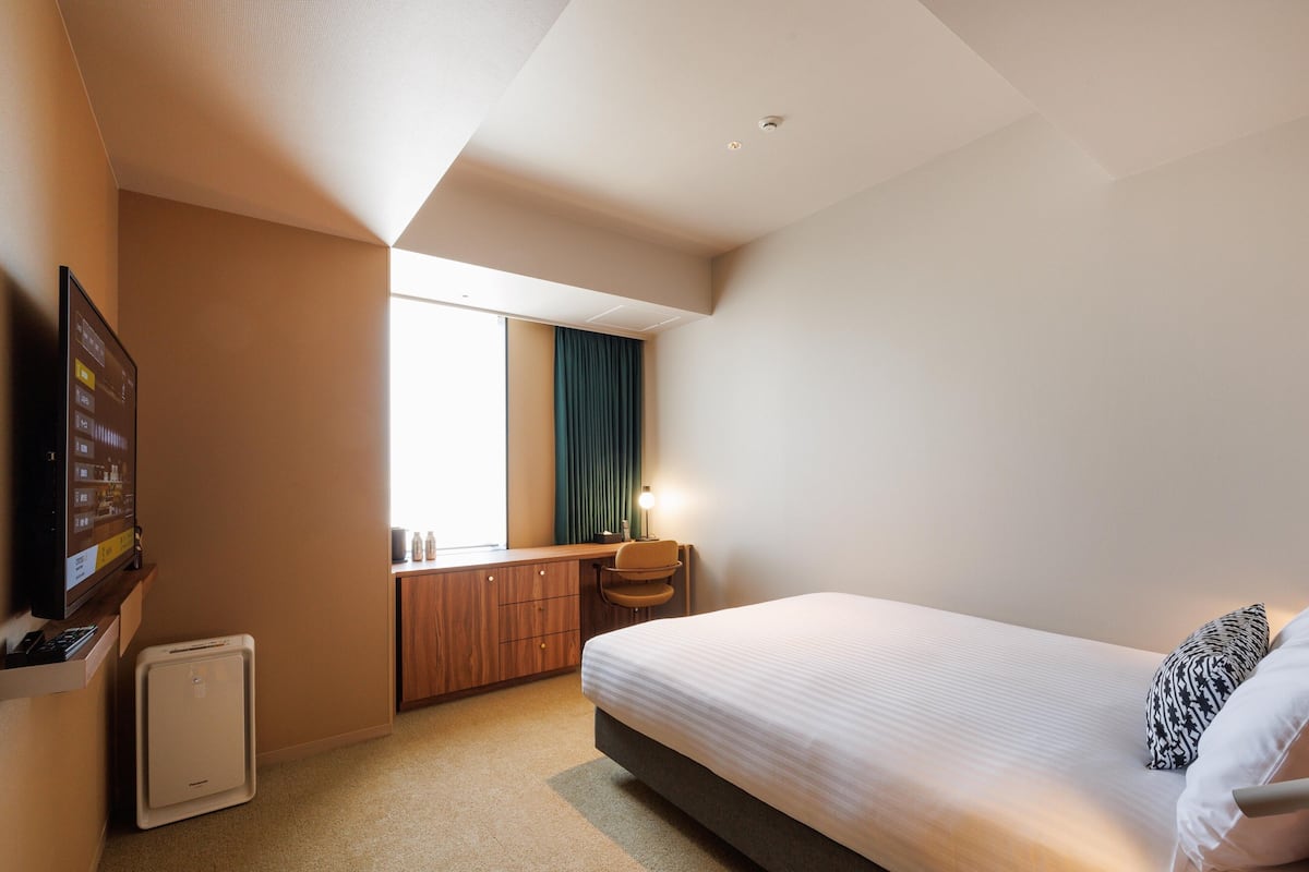 A hotel room with a bed, a desk, a chair, a TV, and a small heater.