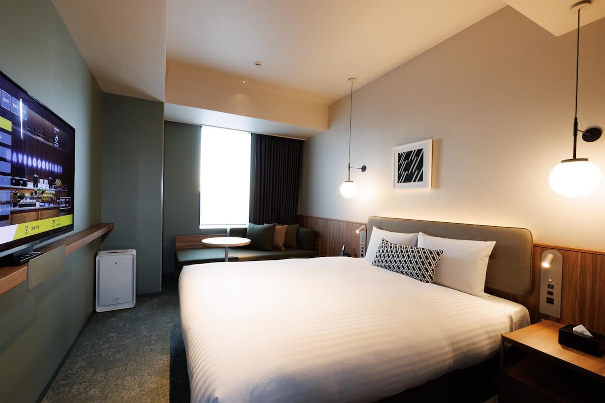 A hotel room with a large bed, a TV on a stand, a small table, and a window with curtains.