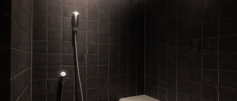 Un área de ducha con loseta oscura, con un banco y un cabezal de ducha manual.