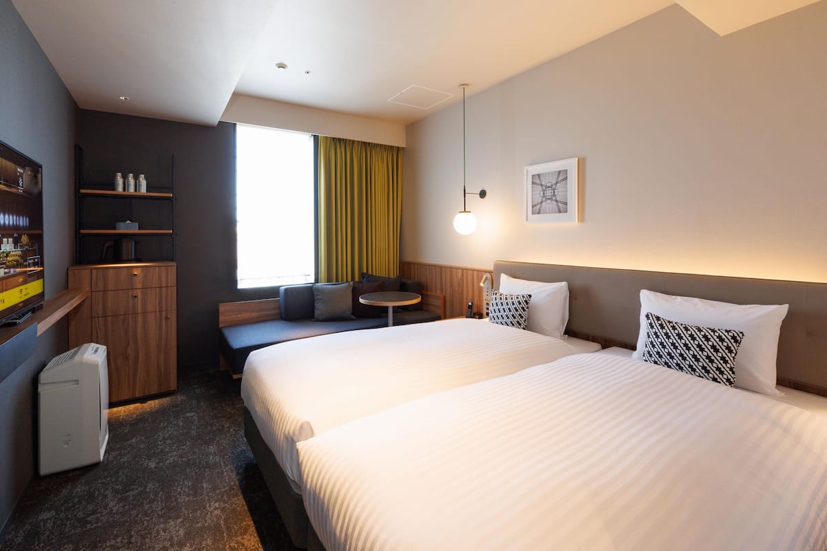A hotel room with two beds, a seating area with a table, a minibar, and a large window with curtains.