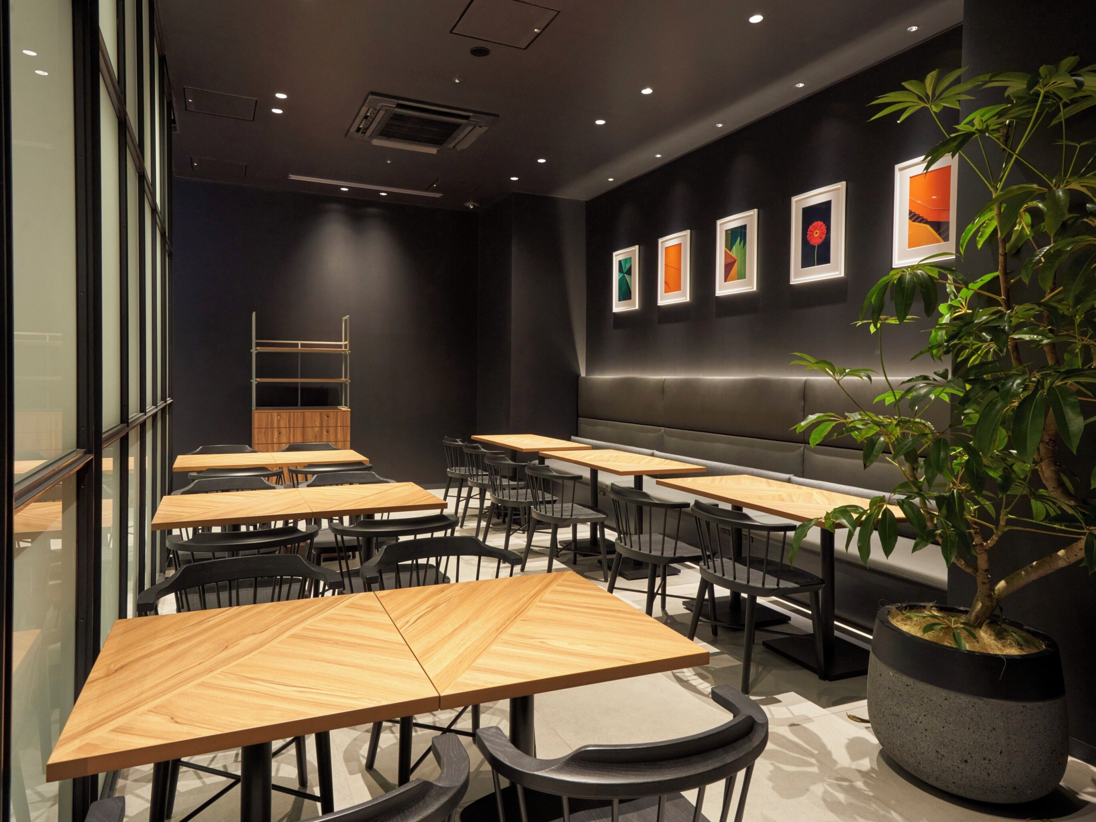 Een modern restaurantinterieur met houten tafels en zwarte stoelen, een zithoek met een bank en een potplant.